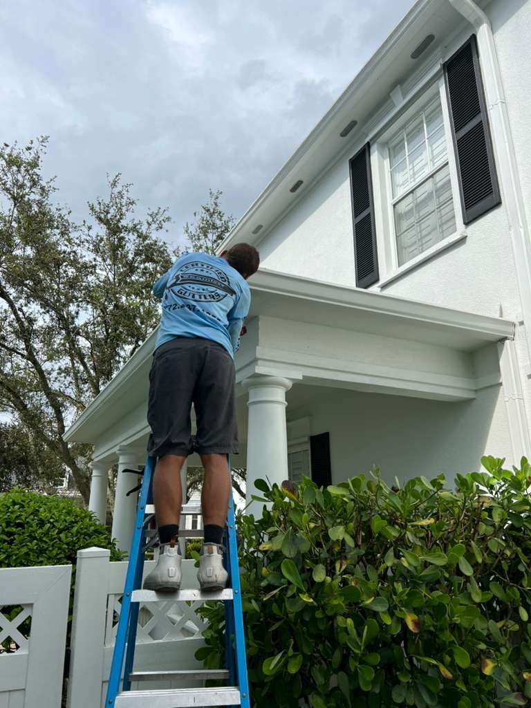 Worker on a ladder servicing gutters at the eave of a two-story white stucco home