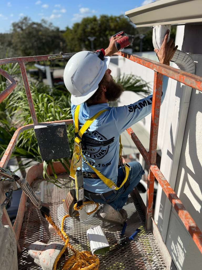 Certified Gutters crew member in a branded shirt and safety harness installing a white downspout from a man-lift on a Florida job site