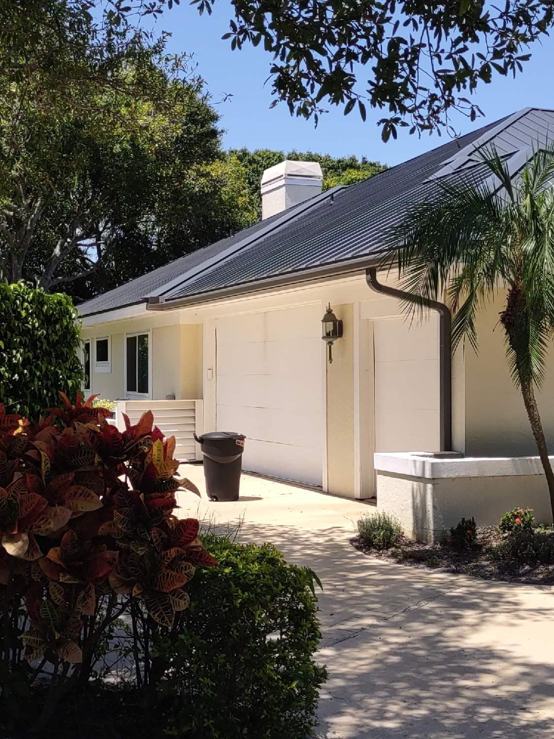 Single-story cream stucco home with a dark metal roof, two-car garage, and dark seamless aluminum gutters and downspouts
