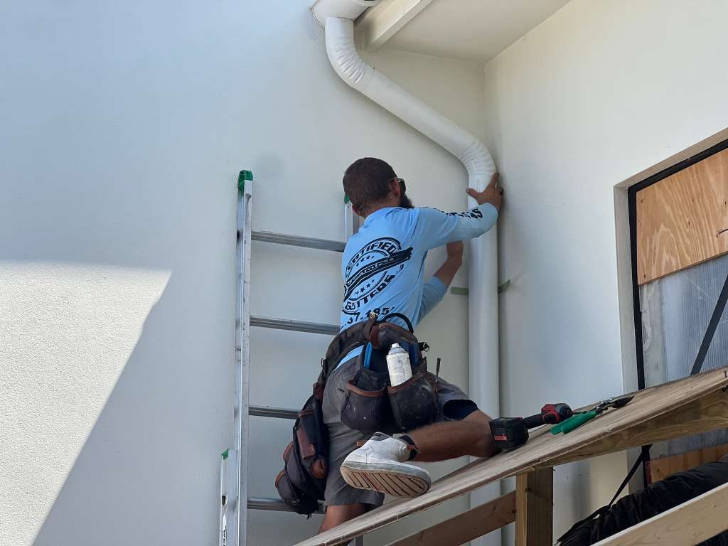 Certified Gutters technician on a ladder installing a white downspout on a stucco home