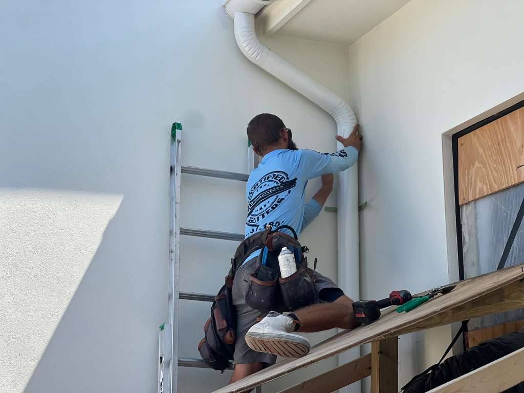 Certified Gutters technician on a ladder installing a white downspout on a stucco home