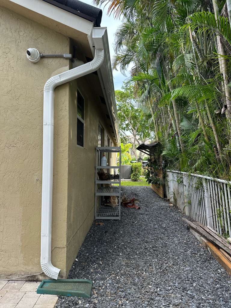 White seamless aluminum gutter and offset downspout running down the side of a tan stucco home with a green splash block and gravel walkway